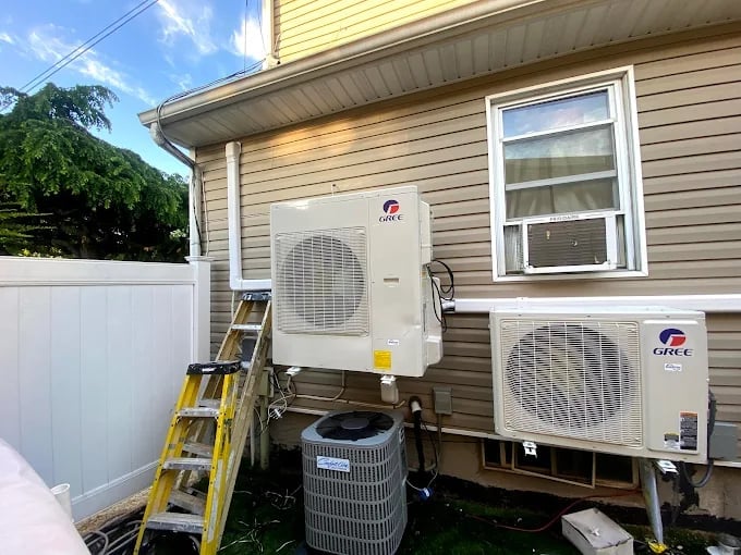Two GREE air conditioning units mounted on a residential house exterior with a yellow ladder and bucket below, beige siding and white window visible