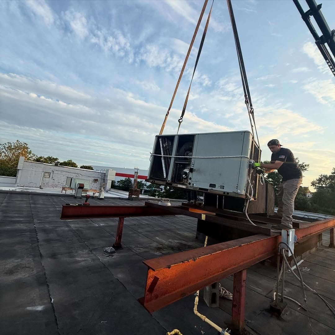Worker uses crane to lift HVAC unit onto rooftop above metal framework with orange and red support beams