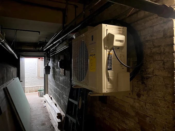 Cluttered basement or utility room with cream-colored air conditioning unit mounted on brick wall and metal piping overhead
