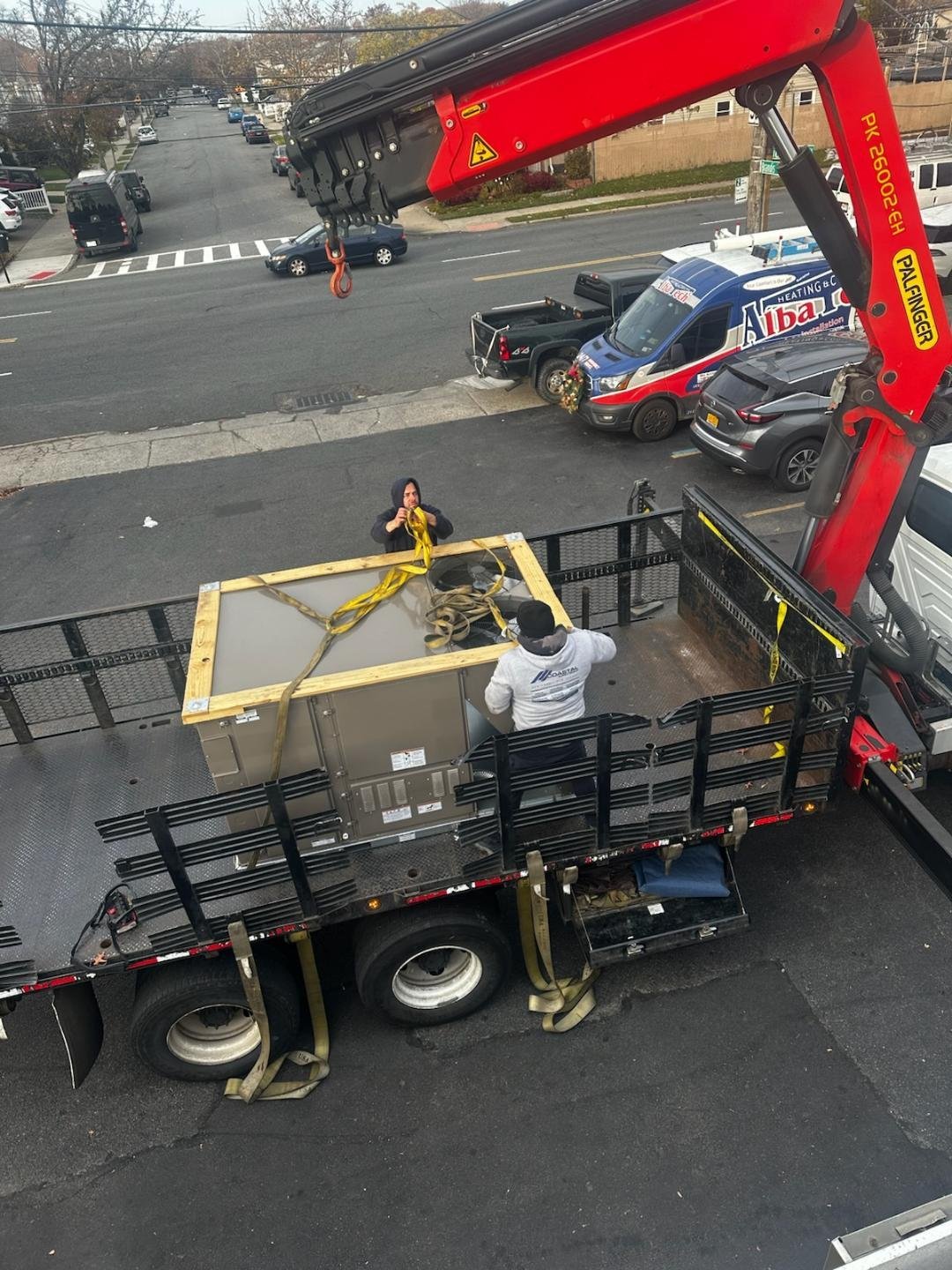 Red and black truck-mounted crane with hydraulic boom lowering yellow pallet of equipment onto flatbed trailer bed