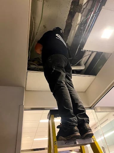 Worker in black clothing standing on yellow ladder, reaching into open ceiling panel with hands above head
