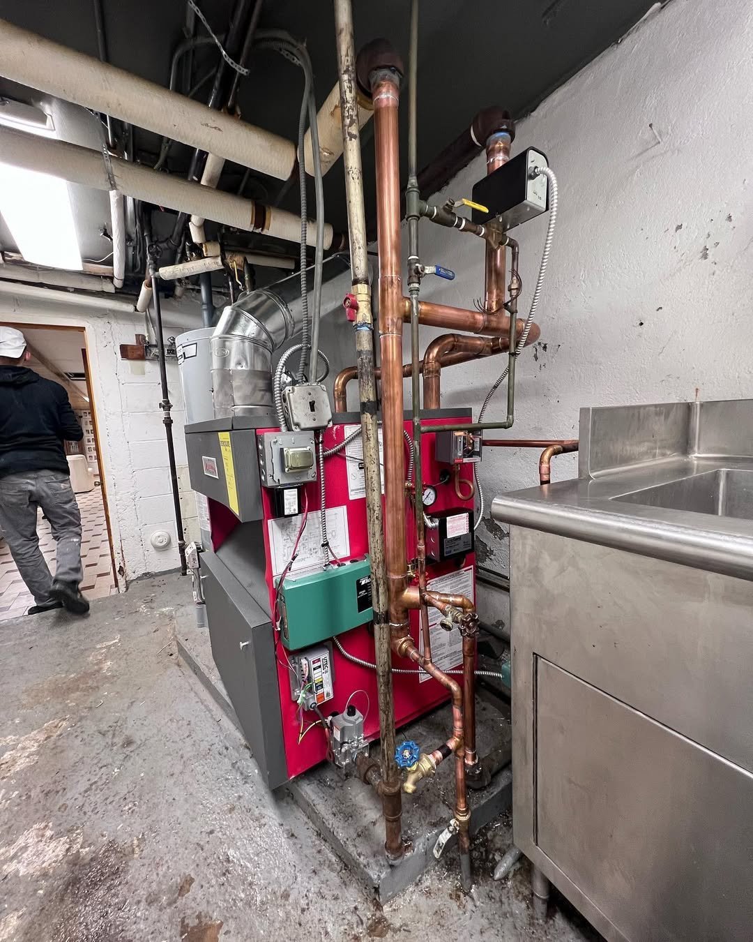 Red and black commercial boiler system with copper pipes in industrial kitchen setting with stainless steel sink