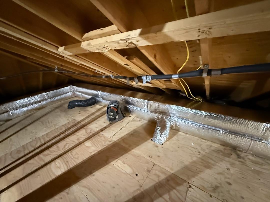 Interior view of wooden attic framing with exposed beams, insulated ductwork, and electrical wiring running across the ceiling and floor