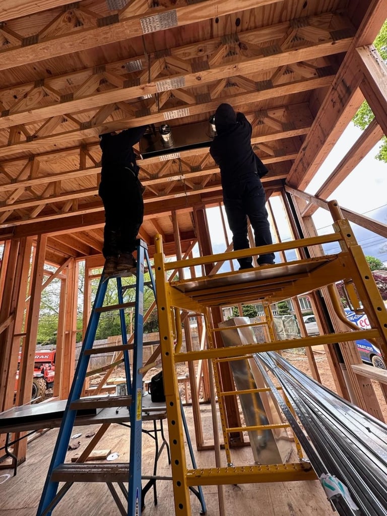 Two workers in black clothing standing on yellow and blue ladders working on wooden roof framing of a residential construction site