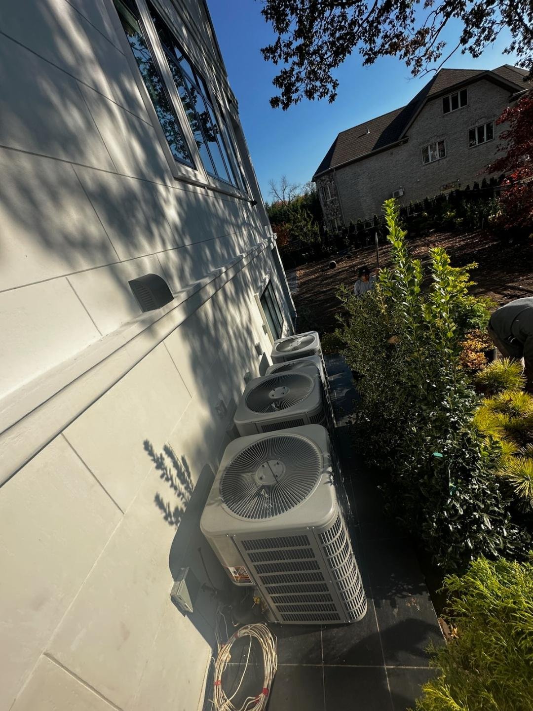 Outdoor HVAC units mounted on white building exterior with stone house and green landscaping visible in background