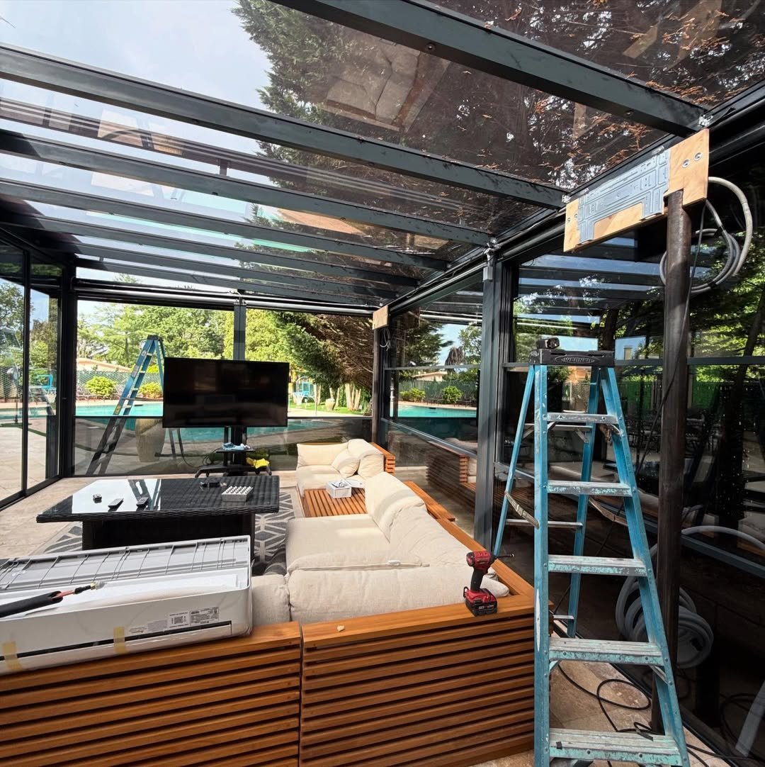 Modern outdoor patio under glass canopy with TV, seating, wooden base, and blue ladder during renovation